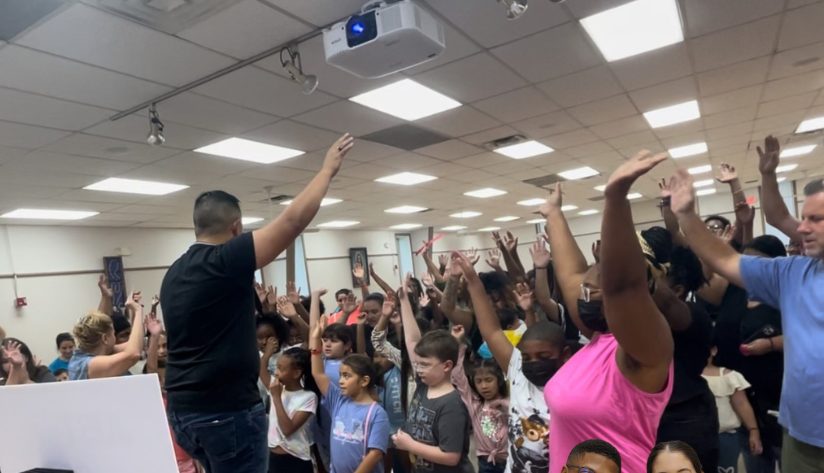 Ace leading a cafeteria full of kids and families with hands raised